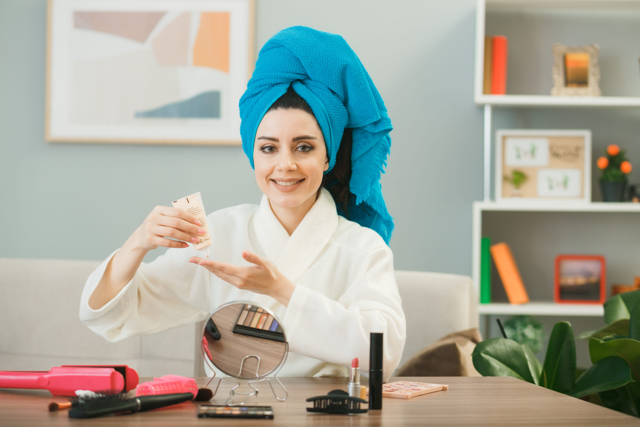 Smiling looking camera young girl holding tone up cream wrapped hair in towel sitting at table with makeup tools in living room
