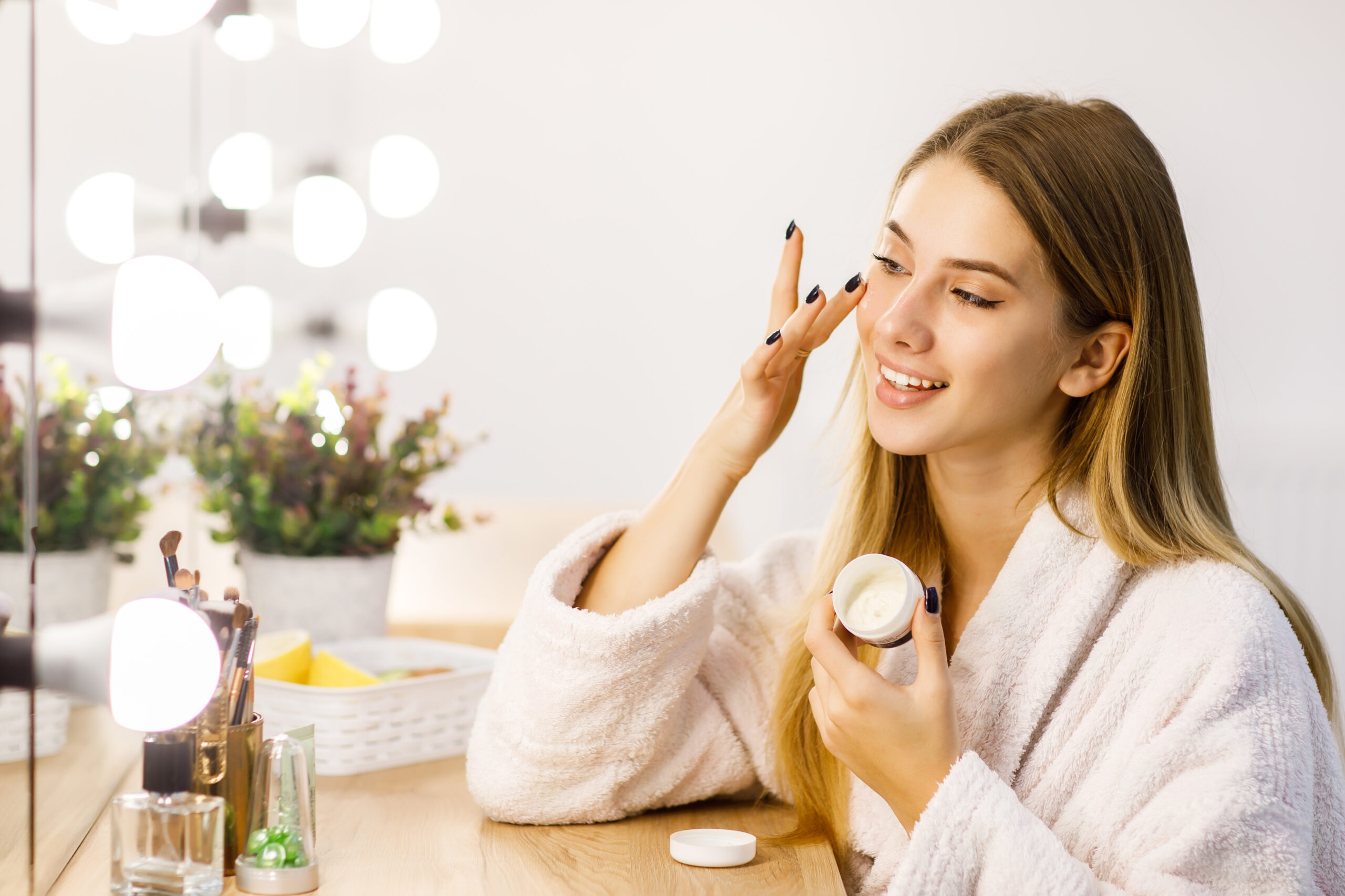 Young beautiful blonde girl cares face skin with moisturizer front mirror